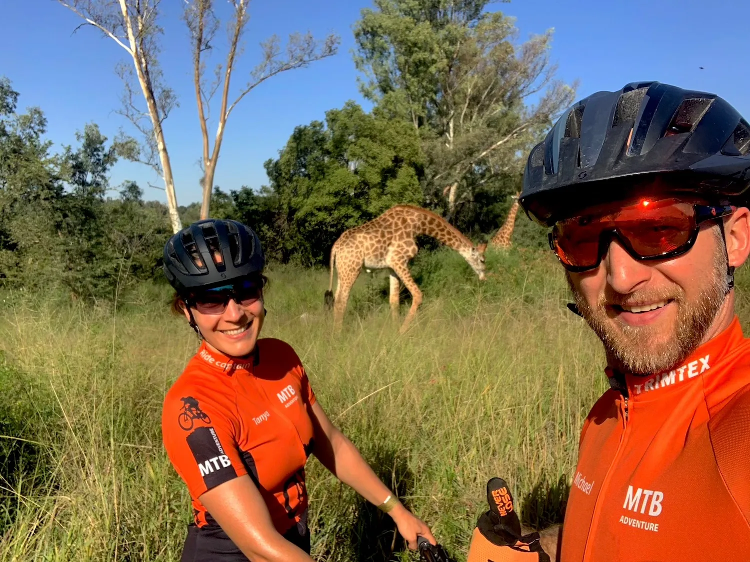 Tanya and Michael Sommer, founders of Gravel Adventure, on a gravel ride in South Africa with giraffes grazing behind