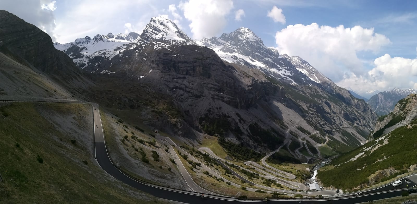 An alpine gravel road climbing through high meadow toward snow-streaked peaks near the Stelvio