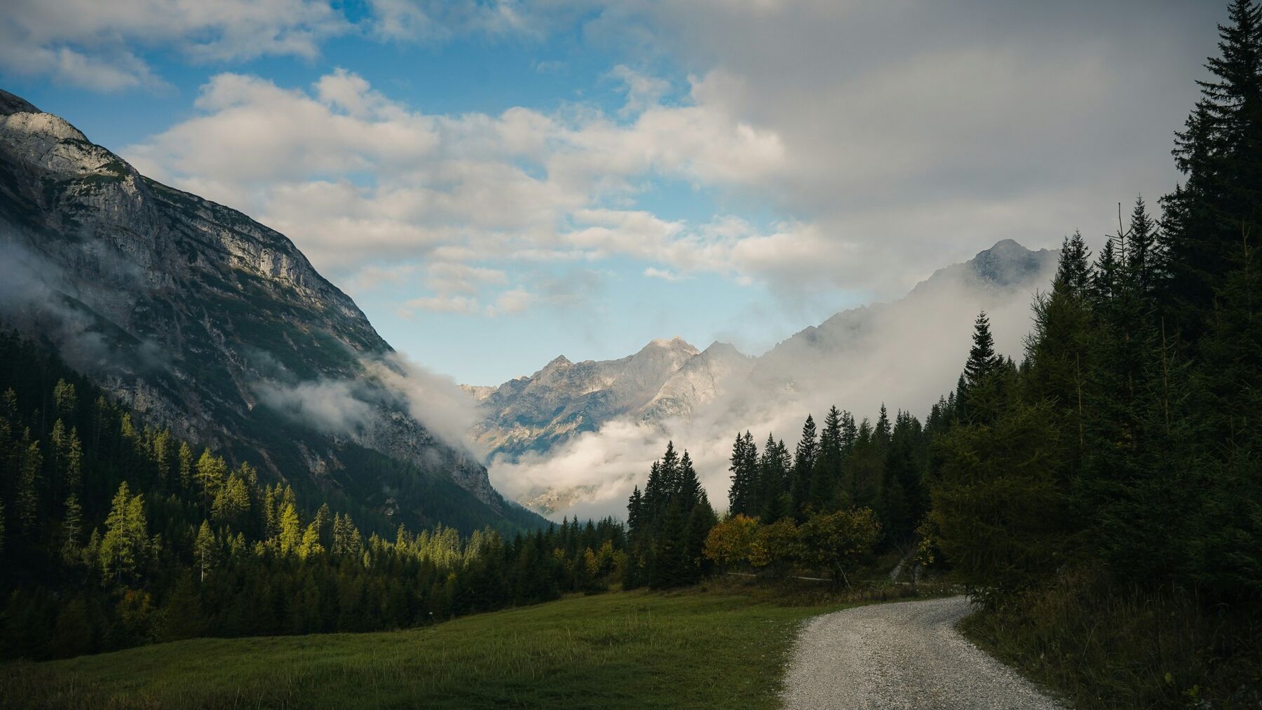 A gravel trail through an alpine valley, mist rolling off the peaks on either side
