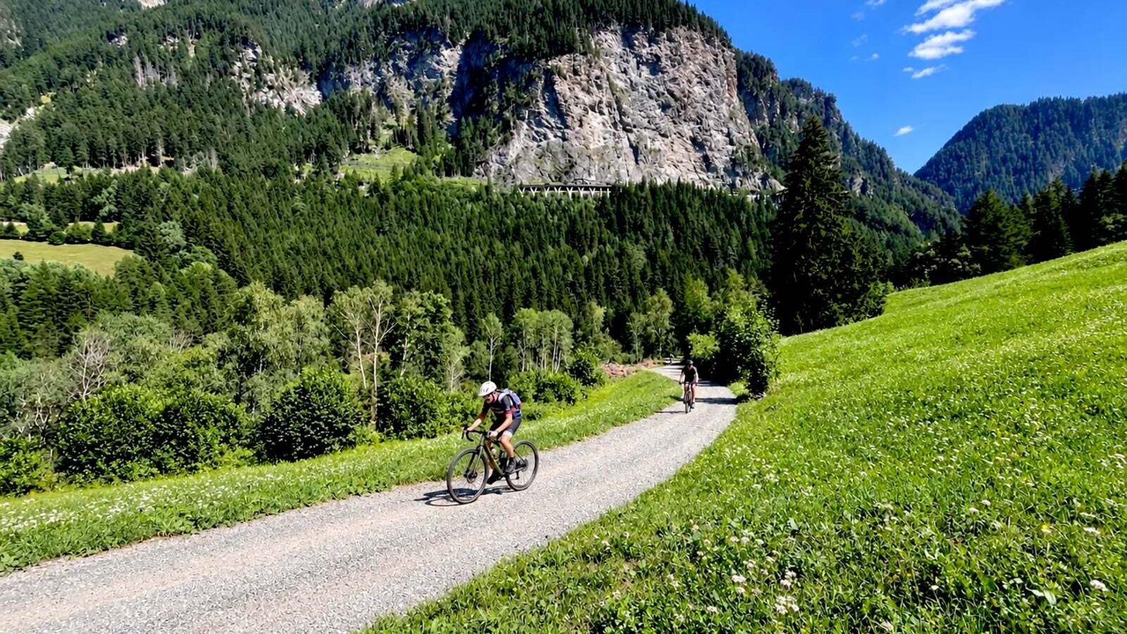 Two gravel cyclists descending a valley gravel road between forested Alpine slopes on the Trans Alp route