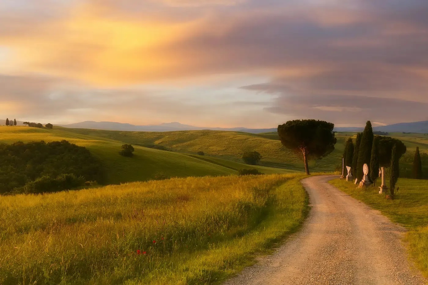 A white gravel road winding between cypresses through Tuscan hills at sunset
